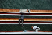 Two individuals are working on a rooftop adorned with rows of green, orange, and red shingles. One person is crouching down while the other is kneeling near a solar panel.