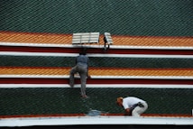 Two individuals are working on a rooftop adorned with rows of green, orange, and red shingles. One person is crouching down while the other is kneeling near a solar panel.
