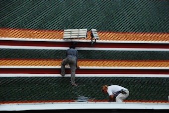 Two individuals are working on a rooftop adorned with rows of green, orange, and red shingles. One person is crouching down while the other is kneeling near a solar panel.