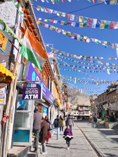 A bustling street scene with colorful prayer flags hanging overhead, people walking along the pavement, and a variety of shops lined with vibrant signage. An ATM is visible with a small queue of people. The backdrop includes a clear blue sky and mountainous landscape.