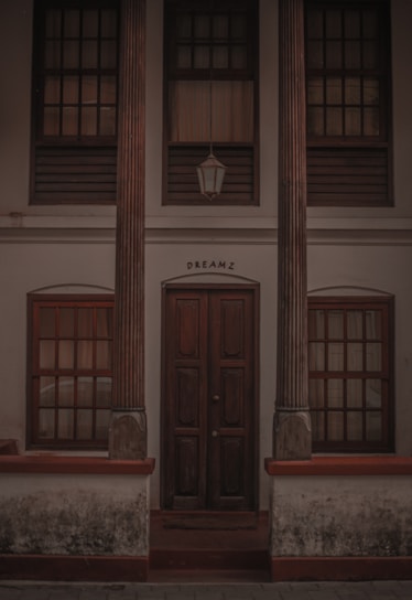 A wooden door is flanked by two tall pillars with intricate carvings. Above the door is a window with wooden shutters and a lantern hanging down. The word 'DREAMZ' is inscribed above the door. The building has a weathered look, with visible signs of age and wear.