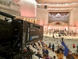 A large indoor auditorium with a grand organ and a setup for an orchestral performance. A professional video camera is positioned on the left side, capturing the scene. Many people are seated in white chairs, waiting for the event to start. The walls and ceiling have elegant architectural designs, and the lighting casts a soft, warm glow throughout the space.