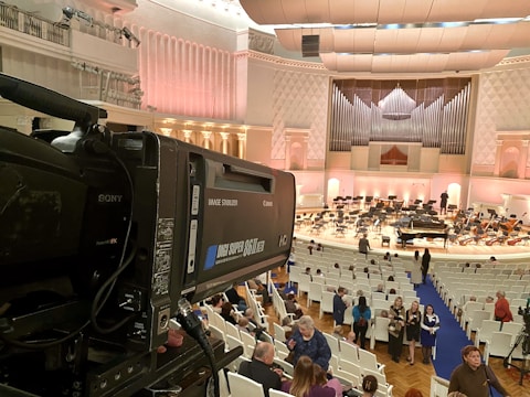 A large indoor auditorium with a grand organ and a setup for an orchestral performance. A professional video camera is positioned on the left side, capturing the scene. Many people are seated in white chairs, waiting for the event to start. The walls and ceiling have elegant architectural designs, and the lighting casts a soft, warm glow throughout the space.