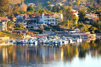 A picturesque waterfront neighborhood featuring several multi-story houses with red-tiled roofs and lush vegetation. Below the houses, numerous boats are docked at a marina, their reflections clearly visible in the calm water. The surrounding area is characterized by rolling hills and an abundance of trees, providing a tranquil and scenic environment.