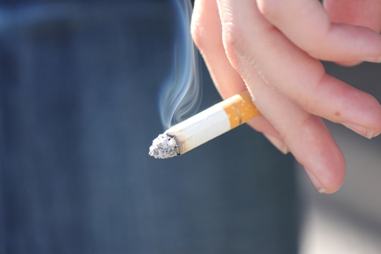 A hand holding a lit cigarette with smoke rising from the tip. The fingers are relaxed, and the background is blurred, focusing attention on the cigarette itself.
