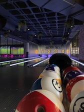 A bowling alley with colorful bowling balls lined up in the foreground. The alley is dimly lit, with number signs '1', '2', '3', '4' lit up in the background. Hanging plants and ceiling lights add to the ambiance.