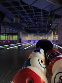A bowling alley with colorful bowling balls lined up in the foreground. The alley is dimly lit, with number signs '1', '2', '3', '4' lit up in the background. Hanging plants and ceiling lights add to the ambiance.