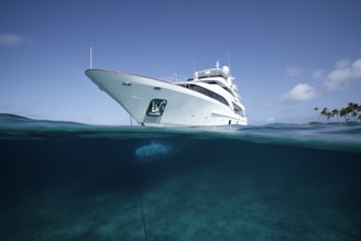 A luxurious white yacht floats on a tranquil blue ocean, partially submerged, with a clear view of its underwater hull. The sky is clear with a few scattered clouds, and a small cluster of palm trees is visible in the distance.
