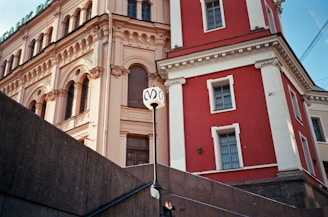 A classical European building with ornate architectural elements in red and beige, featuring arched windows and decorative columns. A street lamp with a distinctive 'M' logo stands in the foreground, and a person is visible on the staircase leading up to the structure.