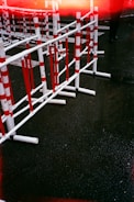 Several red and white striped barriers are positioned on a wet asphalt road. The ground is dark and shiny, indicating recent rain. A partially visible figure in dark clothing is present near the top right corner, and there is a streak of red light across the top of the image.