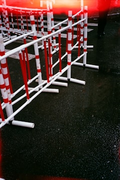 Several red and white striped barriers are positioned on a wet asphalt road. The ground is dark and shiny, indicating recent rain. A partially visible figure in dark clothing is present near the top right corner, and there is a streak of red light across the top of the image.