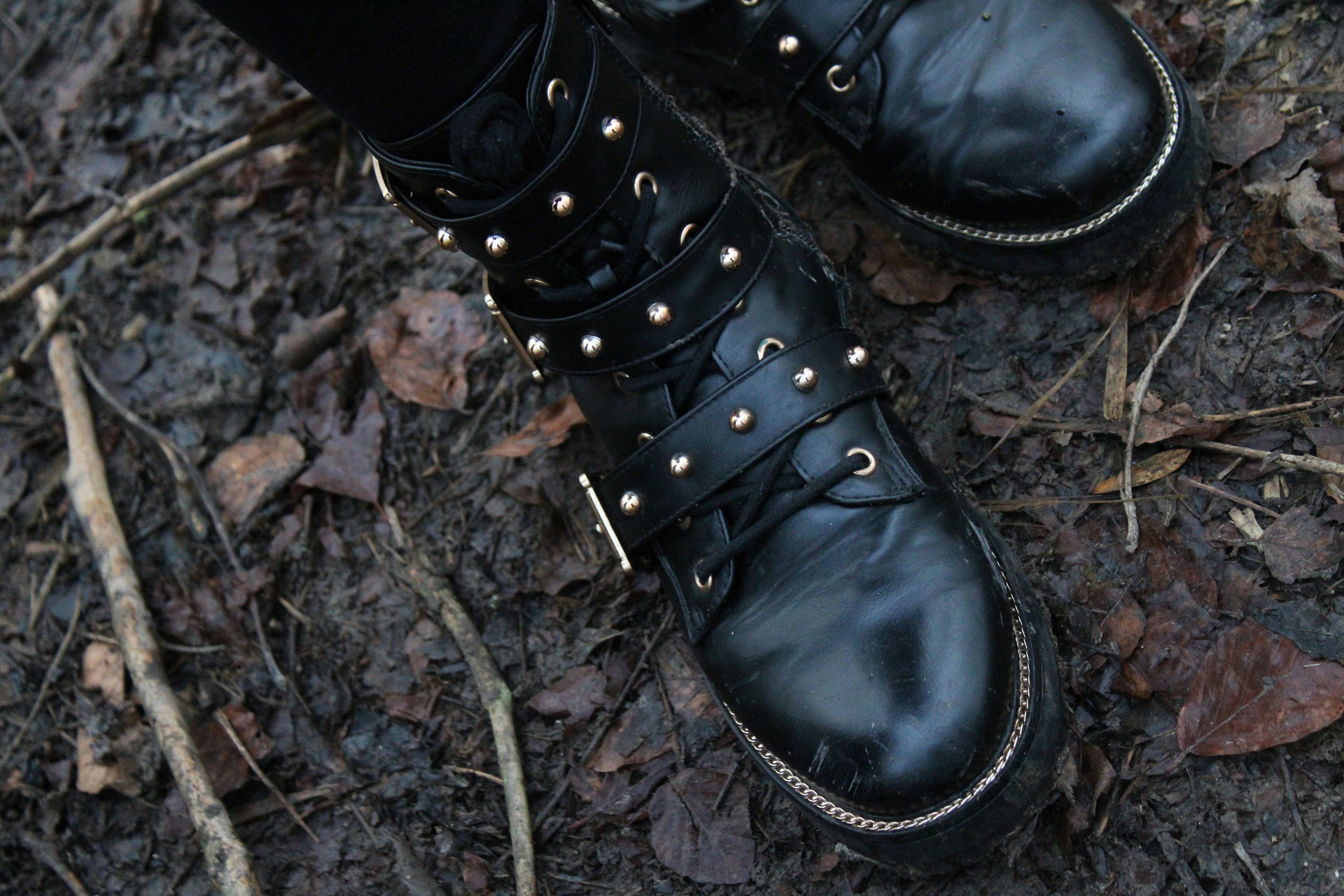A black leather boot with studded straps standing on the muddy, leafy, stick-strewn ground.