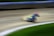 A blurred image capturing a high-speed motorcycle racing around a dirt track. The motion blur emphasizes the speed and dynamic movement of the motorbike, which is positioned at a sharp angle, suggesting a turn. The background includes a blurred track barrier and patches of green, hinting at grass or vegetation alongside the track.