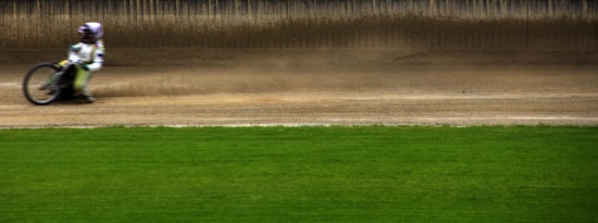 A motorcycle is captured racing across a dirt track with dust trailing behind it. The background consists of a flat dirt surface transitioning to a grassy field in the foreground. The motion blur suggests high speed, and the rider is dressed in protective gear.