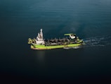 A green and white ship is sailing through calm, dark blue waters. The vessel appears to be equipped with various industrial machinery on its deck, indicating it might be a workboat or a dredger.