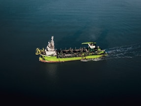 A green and white ship is sailing through calm, dark blue waters. The vessel appears to be equipped with various industrial machinery on its deck, indicating it might be a workboat or a dredger.