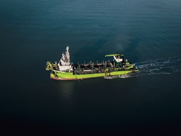 A green and white ship is sailing through calm, dark blue waters. The vessel appears to be equipped with various industrial machinery on its deck, indicating it might be a workboat or a dredger.