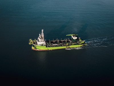A green and white ship is sailing through calm, dark blue waters. The vessel appears to be equipped with various industrial machinery on its deck, indicating it might be a workboat or a dredger.