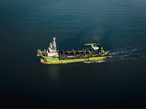 A green and white ship is sailing through calm, dark blue waters. The vessel appears to be equipped with various industrial machinery on its deck, indicating it might be a workboat or a dredger.
