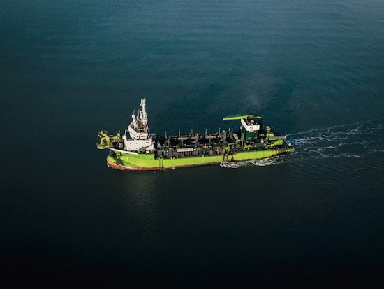 A green and white ship is sailing through calm, dark blue waters. The vessel appears to be equipped with various industrial machinery on its deck, indicating it might be a workboat or a dredger.