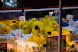 A collection of plush toys inside a claw machine is visible, with several yellow Pikachu dolls prominently displayed. The toys are lying on a pile of white stuffing material, and there are other plush animals in different colors, including a white and yellow bear dressed as a chef.