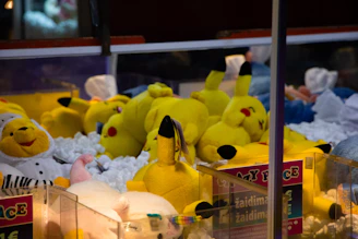 A collection of plush toys inside a claw machine is visible, with several yellow Pikachu dolls prominently displayed. The toys are lying on a pile of white stuffing material, and there are other plush animals in different colors, including a white and yellow bear dressed as a chef.
