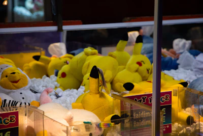 A collection of plush toys inside a claw machine is visible, with several yellow Pikachu dolls prominently displayed. The toys are lying on a pile of white stuffing material, and there are other plush animals in different colors, including a white and yellow bear dressed as a chef.