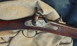 A close-up view of a vintage rifle, specifically a U.S. Springfield from 1861. The rifle's metal components are set against a wooden stock with a rich brown hue. It rests on a textured, light beige fabric.