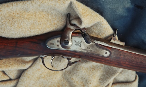 A close-up view of a vintage rifle, specifically a U.S. Springfield from 1861. The rifle's metal components are set against a wooden stock with a rich brown hue. It rests on a textured, light beige fabric.