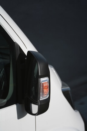 A close-up view of a vehicle's side mirror, featuring an integrated indicator light with a black casing. The white body of the vehicle contrasts against the dark background.
