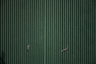 A close-up view of a corrugated metal surface with vertical ridges, painted in a muted green color. There are some signs of wear and small pieces of tape or paint chips present on the surface.