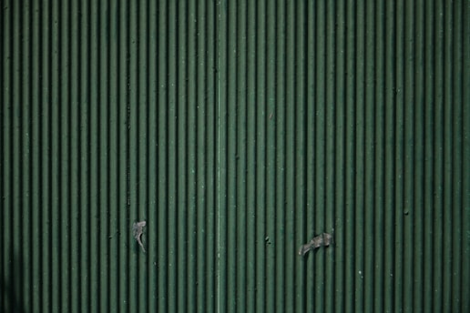 A close-up view of a corrugated metal surface with vertical ridges, painted in a muted green color. There are some signs of wear and small pieces of tape or paint chips present on the surface.