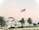 A large white coastal house with a traditional design, surrounded by palm trees. An American flag is prominently displayed on a tall flagpole in the foreground. The sky is a pastel blend of pink and blue, suggesting early morning or late afternoon.