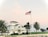 A large white coastal house with a traditional design, surrounded by palm trees. An American flag is prominently displayed on a tall flagpole in the foreground. The sky is a pastel blend of pink and blue, suggesting early morning or late afternoon.
