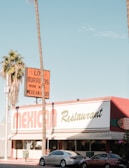 A Mexican restaurant with a vintage-style sign that reads 'Los Burritos Home Made Mexican Food.' The building is red and white with green accents. Two cars are parked in front, and tall palm trees are visible on either side of the image against a clear blue sky.
