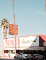 A Mexican restaurant with a vintage-style sign that reads 'Los Burritos Home Made Mexican Food.' The building is red and white with green accents. Two cars are parked in front, and tall palm trees are visible on either side of the image against a clear blue sky.