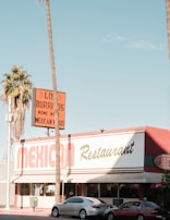 A Mexican restaurant with a vintage-style sign that reads 'Los Burritos Home Made Mexican Food.' The building is red and white with green accents. Two cars are parked in front, and tall palm trees are visible on either side of the image against a clear blue sky.