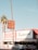 A Mexican restaurant with a vintage-style sign that reads 'Los Burritos Home Made Mexican Food.' The building is red and white with green accents. Two cars are parked in front, and tall palm trees are visible on either side of the image against a clear blue sky.