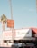 A Mexican restaurant with a vintage-style sign that reads 'Los Burritos Home Made Mexican Food.' The building is red and white with green accents. Two cars are parked in front, and tall palm trees are visible on either side of the image against a clear blue sky.