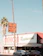 A Mexican restaurant with a vintage-style sign that reads 'Los Burritos Home Made Mexican Food.' The building is red and white with green accents. Two cars are parked in front, and tall palm trees are visible on either side of the image against a clear blue sky.