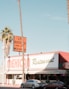 A Mexican restaurant with a vintage-style sign that reads 'Los Burritos Home Made Mexican Food.' The building is red and white with green accents. Two cars are parked in front, and tall palm trees are visible on either side of the image against a clear blue sky.