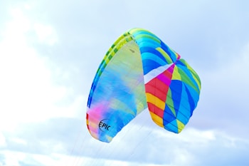 A colorful, multicolored parachute with vivid stripes of blue, teal, red, yellow, and green billows against a cloudy sky. The fabric appears to be made of translucent material, allowing light to pass through it, enhancing its vibrancy. The word 'EPIC' is visible on one section of the parachute.