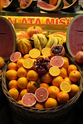 A variety of fruits neatly arranged at a market stall. The centerpiece features a large basket filled with fresh oranges, decorated with halves of oranges, grapefruits, and a pomegranate. Surrounding the basket, there are bananas, small pumpkins, and watermelons with their vibrant red flesh visible.