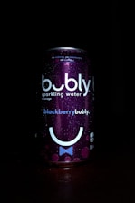 A can of bubly sparkling water is prominently featured against a dark background. The can has a deep purple color with the label 'blackberrybubly'. It includes bubbles and a stylized smiley face design. Water droplets are visible on the surface, suggesting condensation.