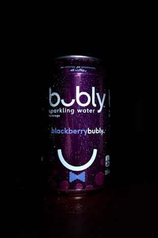 A can of bubly sparkling water is prominently featured against a dark background. The can has a deep purple color with the label 'blackberrybubly'. It includes bubbles and a stylized smiley face design. Water droplets are visible on the surface, suggesting condensation.