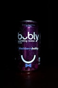 A can of bubly sparkling water is prominently featured against a dark background. The can has a deep purple color with the label 'blackberrybubly'. It includes bubbles and a stylized smiley face design. Water droplets are visible on the surface, suggesting condensation.