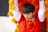 A person wearing a red costume with intricate gold designs, holding up what appears to be a piece of a colorful, textured lion dance costume. Bright orange and yellow fabric surrounds the individual, suggesting movement and festivities.