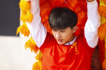 A person wearing a red costume with intricate gold designs, holding up what appears to be a piece of a colorful, textured lion dance costume. Bright orange and yellow fabric surrounds the individual, suggesting movement and festivities.