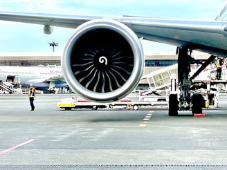 A close-up view of a large aircraft jet engine on a runway with a terminal in the background. An airport worker in a reflective vest and helmet is visible nearby, along with other airport equipment and vehicles. Another plane is parked in the distance.