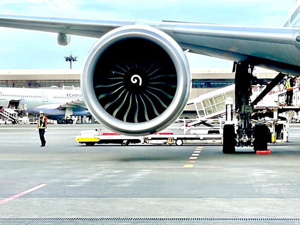 A close-up view of a large aircraft jet engine on a runway with a terminal in the background. An airport worker in a reflective vest and helmet is visible nearby, along with other airport equipment and vehicles. Another plane is parked in the distance.
